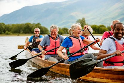 A group of older friends laugh as they row a long, wooden canoe.