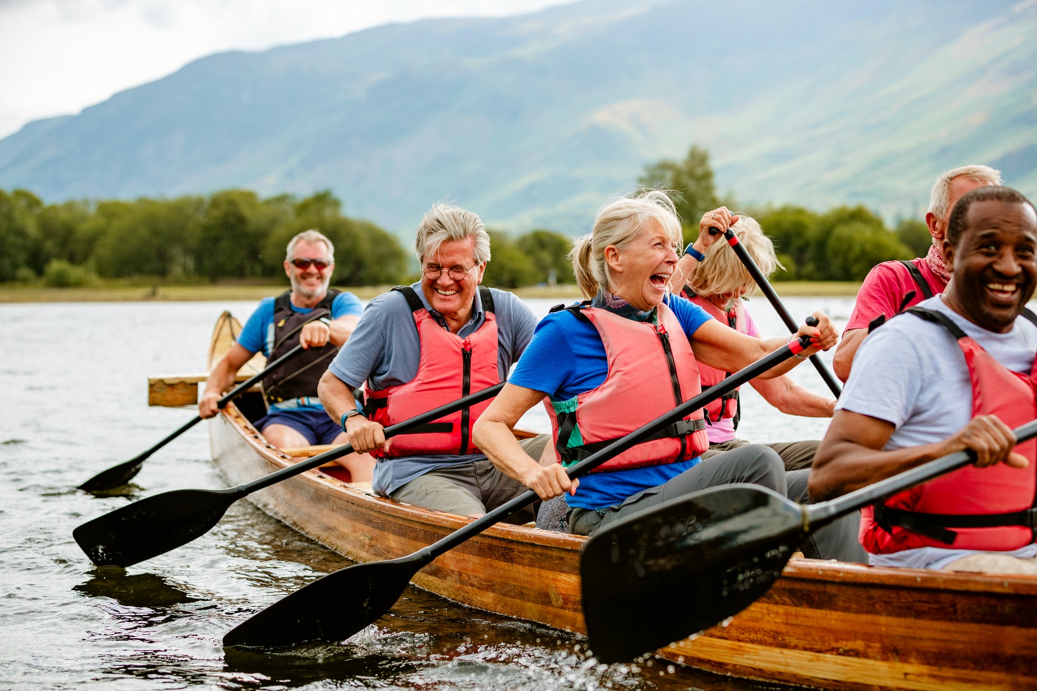 A group of older friends laugh as they row a long, wooden canoe.