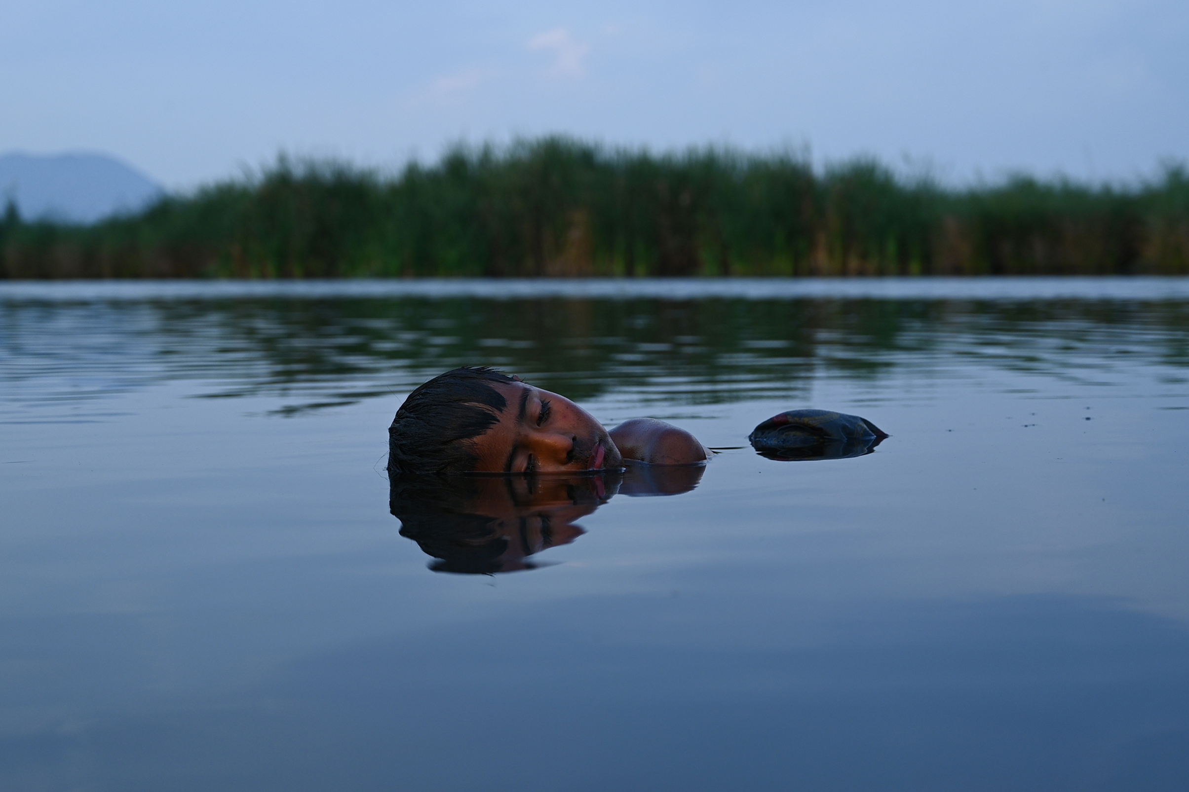 A child peacefully floats face-up in calm water, eyes closed, with reeds in the background and a serene, tranquil atmosphere at dusk