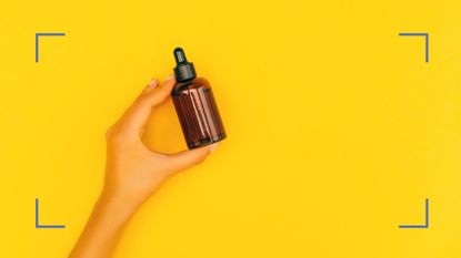 Bottle of argan oil being held by a hand against a yellow background. Blue illustrative corner details in each corner 