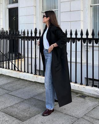 influencer Anna Newton poses on a London sidewalk wearing a long black coat, white tee, straight-leg jeans, and loafers