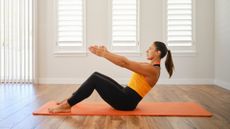 woman wearing an orange vest and black leggings performing a pilates roll-up movement from a seated position on an orange yoga mat with white windows and walls behind her.