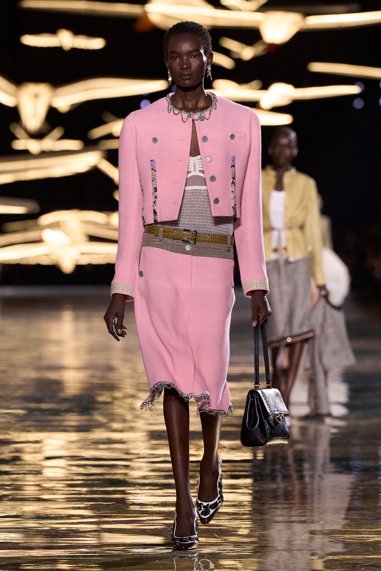 A Chanel spring/summer 2027 runway model wearing a pink tweed jacket styled with a matching pencil skirt, sheer top, brown belt, and pumps.