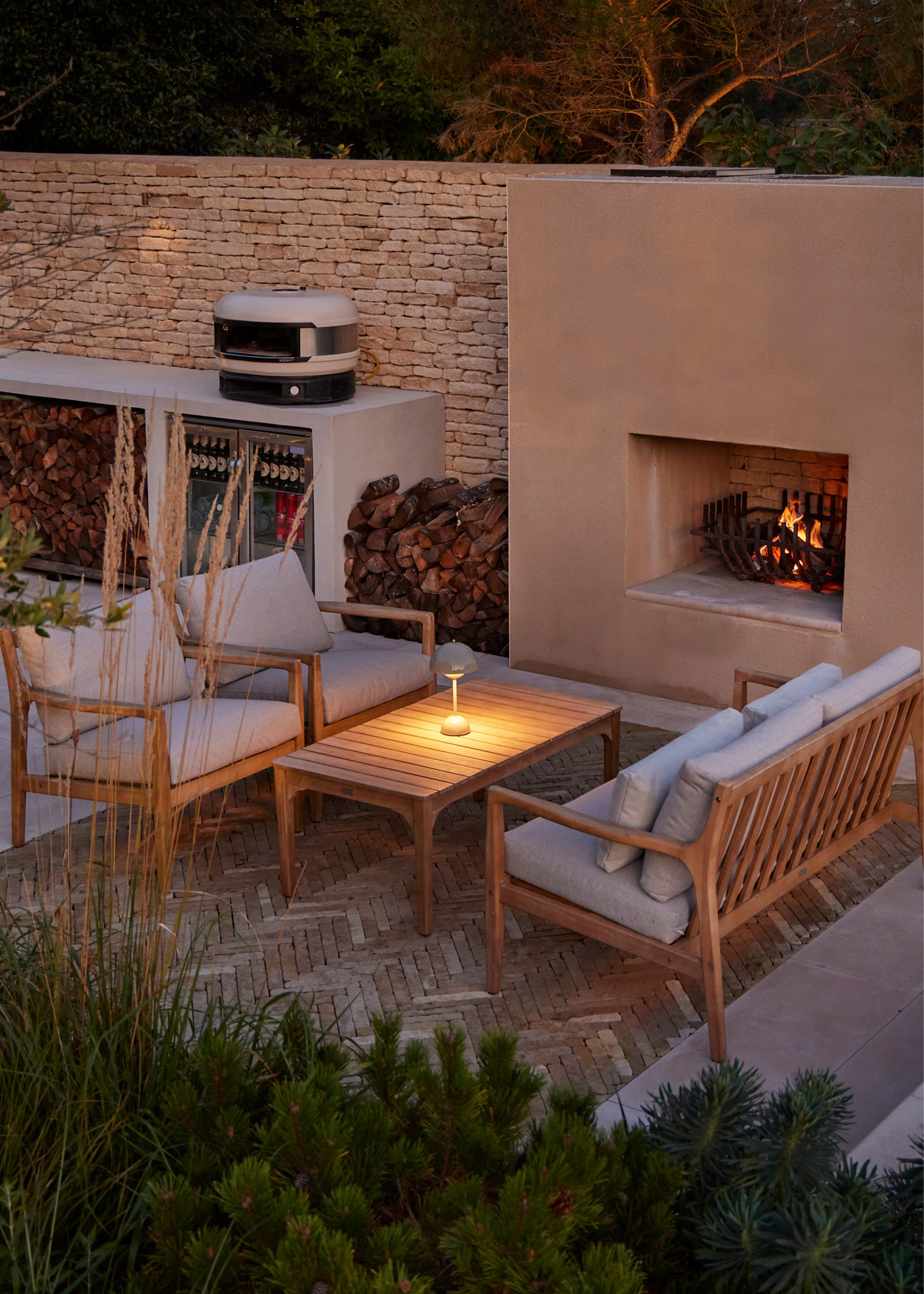 An outdoor seating area that has two garden sofas facing each other with a coffee table in the center with a lit portable lamp on top, next to a lit fireplace by an outdoor kitchen with a pizza oven