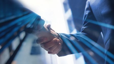 Two businessmen shake hands against backdrop of blurred office buildings.