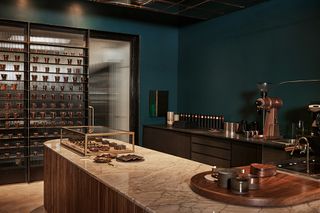 Inerior of the store featuring dark green walls and beige concrete floor. Display of Milla Chocolates hazelnut coffee truffles on multiple levels shelf against blurred glass. A display of bon bons on a marble top island with wood base.
