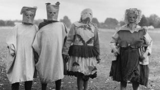 Four children stand outside in a field wearing eerie, homemade Halloween costumes, with two wearing paper bags over their heads and the others wearing grotesque-looking masks.