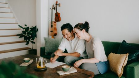A happy couple in their late forties relax on a couch, drinking tea from Japanese cups. A guitar and ukulele hang on the wall behind them.
