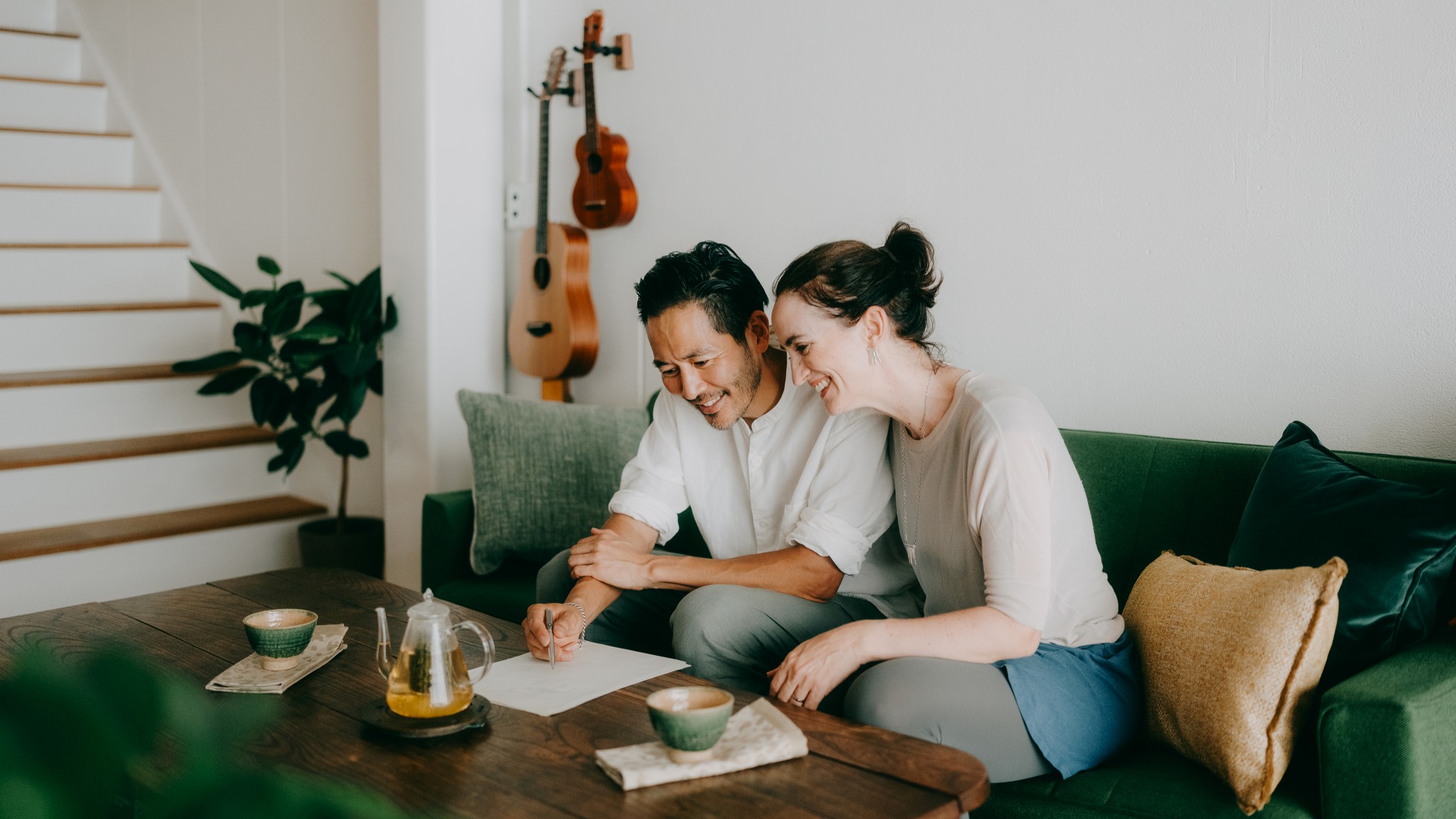 A happy couple in their late forties relax on a couch, drinking tea from Japanese cups. A guitar and ukulele hang on the wall behind them.