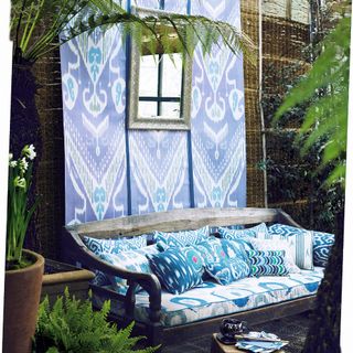 souk style garden theme with blue patterned cushions and fabric behind bench, silver patterned mirror on fabric. Bench with cushions and upholstered seating