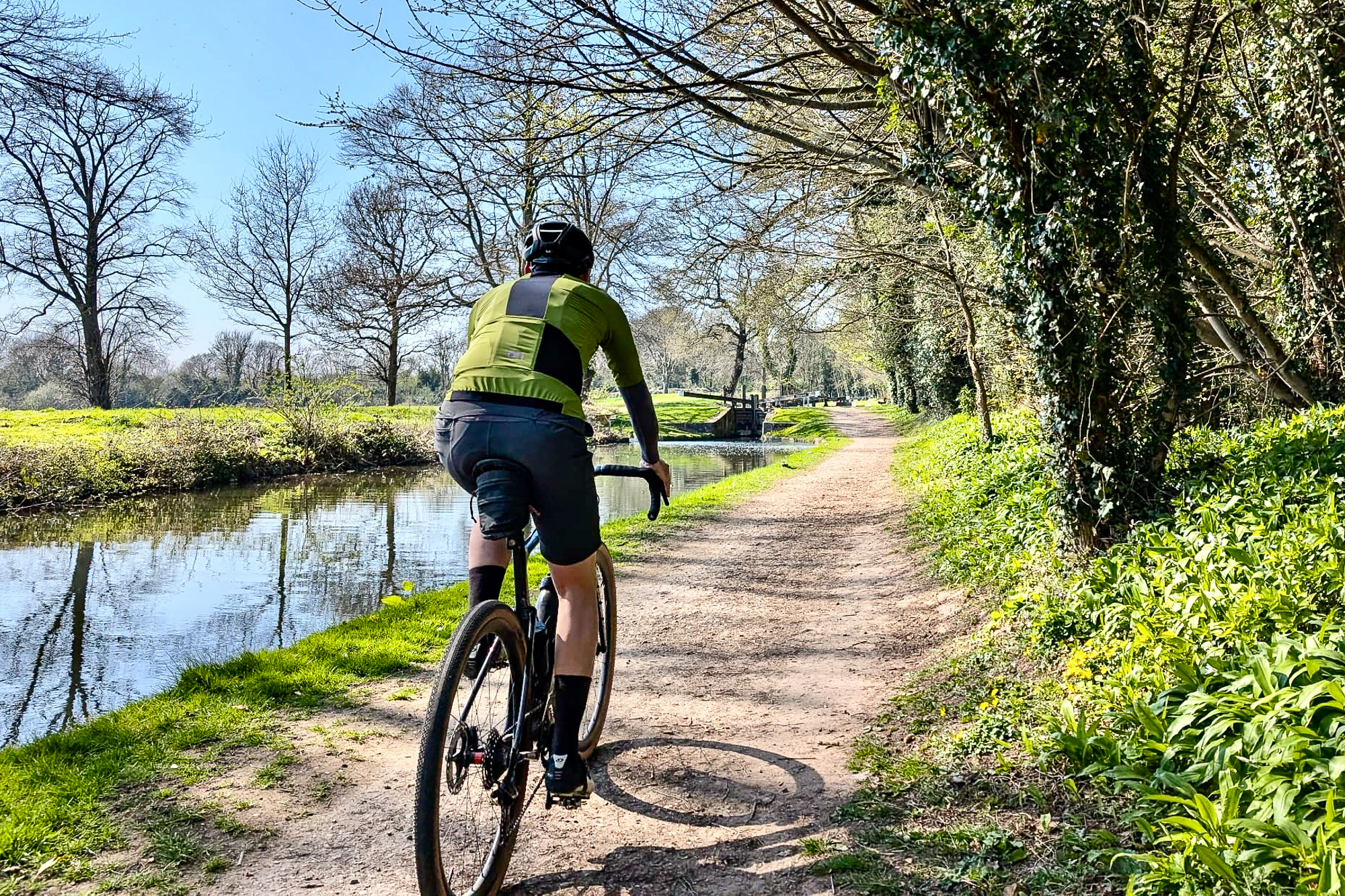 man riding away from the camera wearing a green jersey along a canal towpath