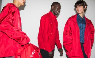 Three male models wearing looks from Off-White's collection. One model is wearing a red jacket and is holding a red bag with white wording. Another model is wearing a red shirt, red jacket and black trousers. And the third model is wearing a blue denim shirt, red jacket and dark coloured trousers