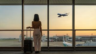 woman with suitcase looking outside airport window at plane taking off