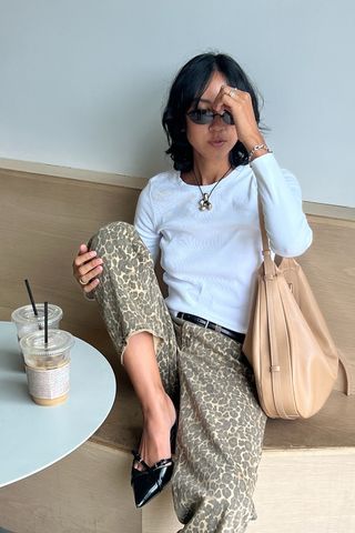 Eine Frau sitzt in einem Café und trägt ein weißes T-Shirt mit schwarzem Gürtel, Leopardenjeans, schwarze spitze Kitten-Heels und eine lässige braune Tasche.