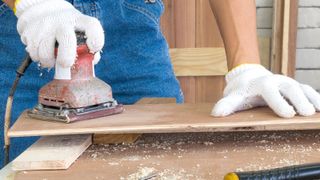 Man with white gloves holding a well used red orbital sander, sanding a sheet of plywood on a wooden bench
