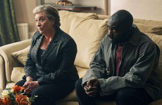 Wes (Caroline Quentin) and George (Paapa Essiedu) sit on a soft stone-hued sofa. They are both looking up at a third, unseen, person.