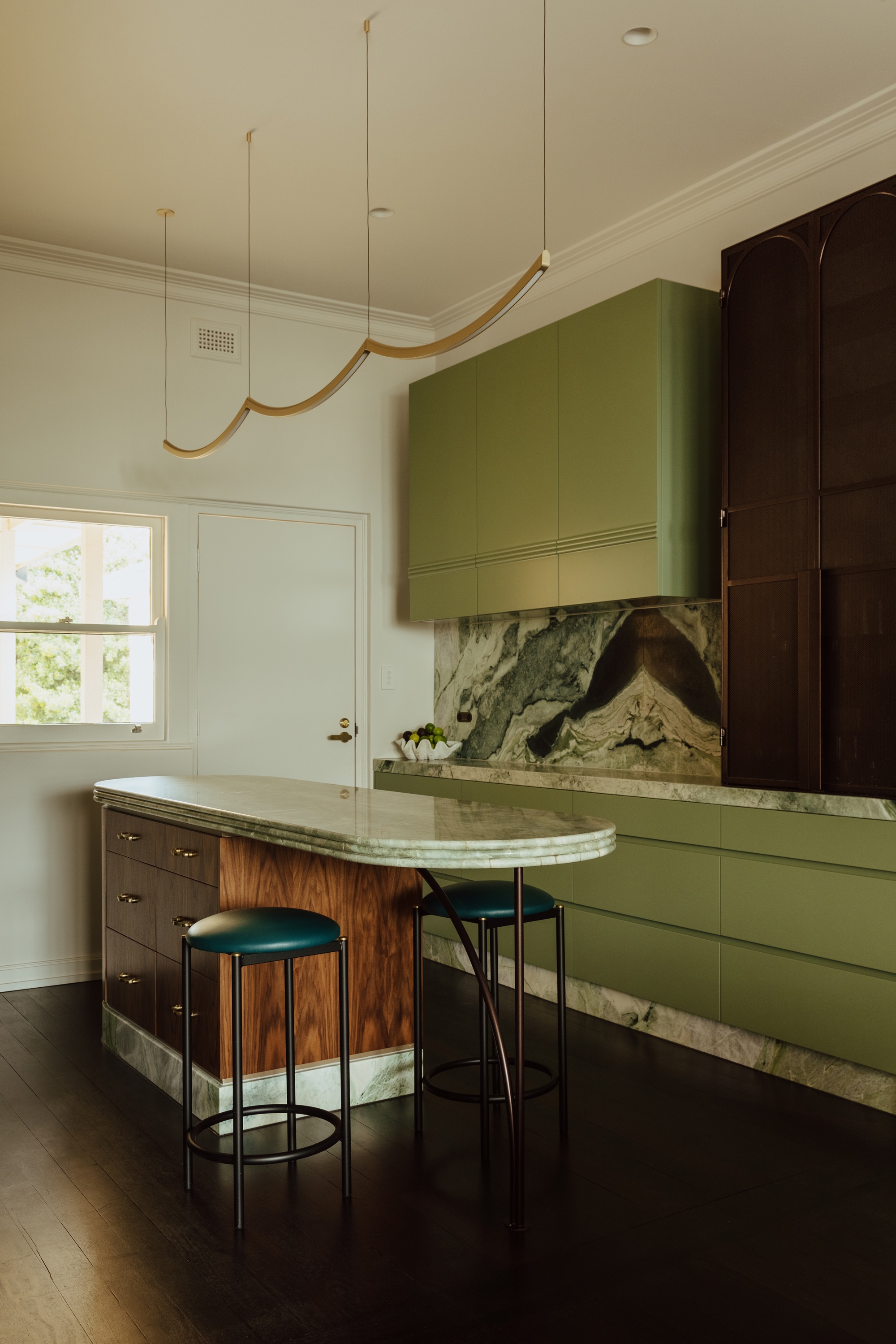 a small green kitchen with an art deco inspired styling, the island is curved with a detailed profile counter and a burl wood base. the backsplash is a rich marbled green natural stone