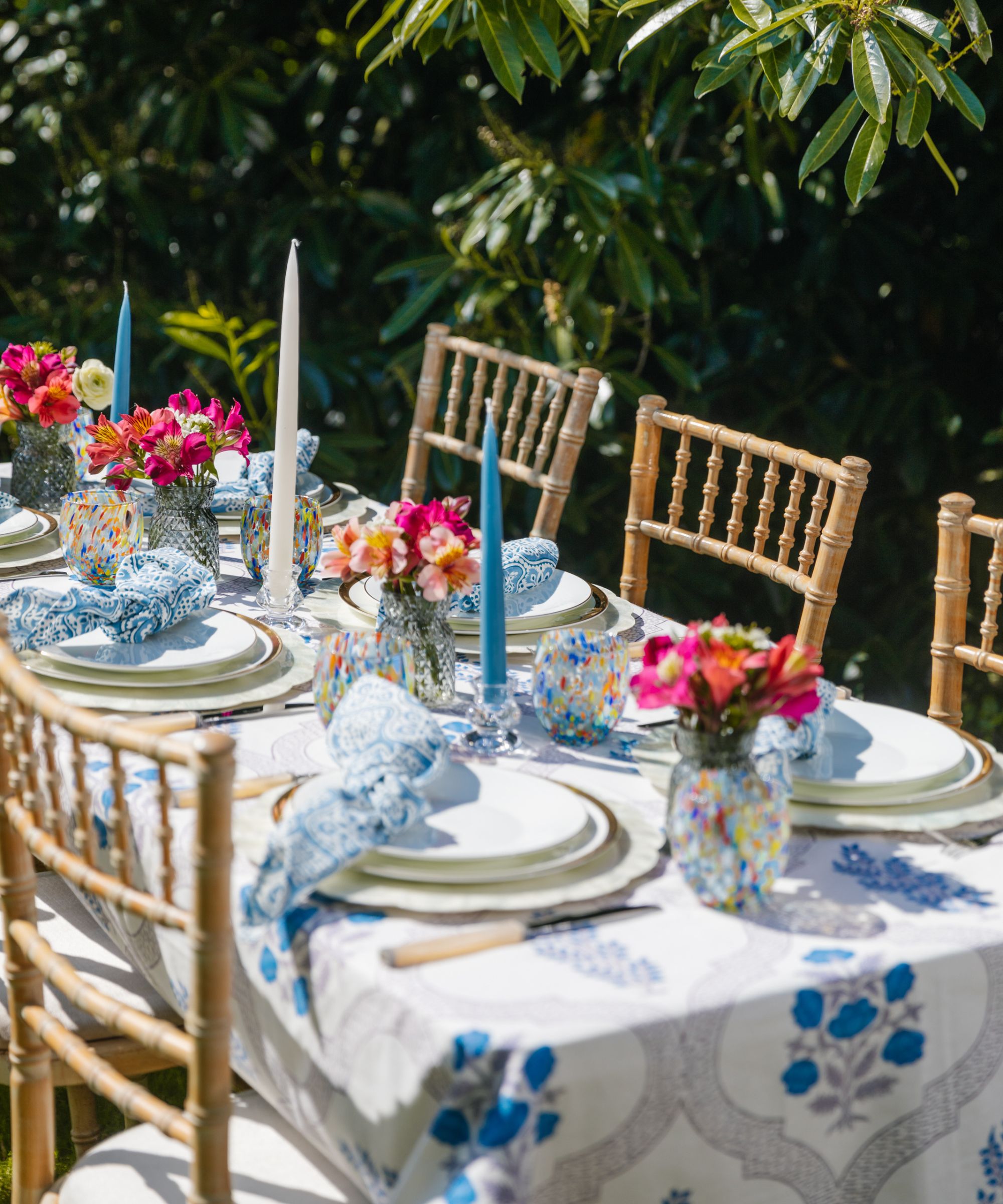 outdoor dining table with bamboo style chairs with a blue and white floral tablecloth, white plates, and tall blue tape candles and small floral bud vases