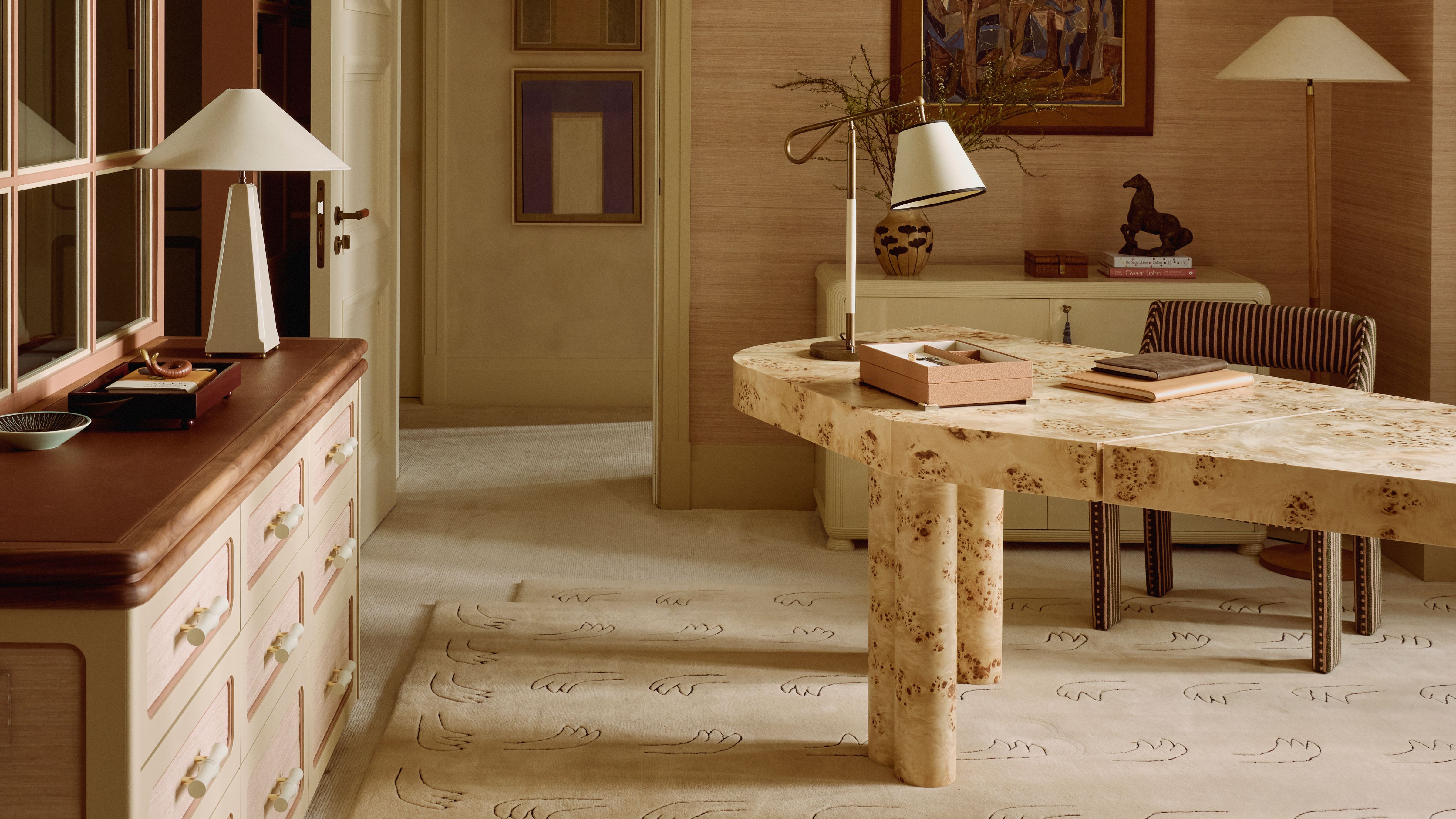 Neutral office space featuring an asymmetrical burl wood desk, linen-shaded table lamps, wall art, a figural motif beige rug, and a striped office chair.