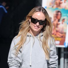 Olivia Wilde wearing a gray hoodie, black trousers, black pointed-toe shoes, and black sunglasses