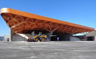 Daytime, external image of the salt storage at the Highway Support Centre, Assen, triangular orange roof, concrete area around the structure, yellow tractor, man walking, clear blue sky