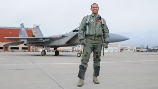a man in a military pilot flight suit posing in front of a jet aircraft and hangars