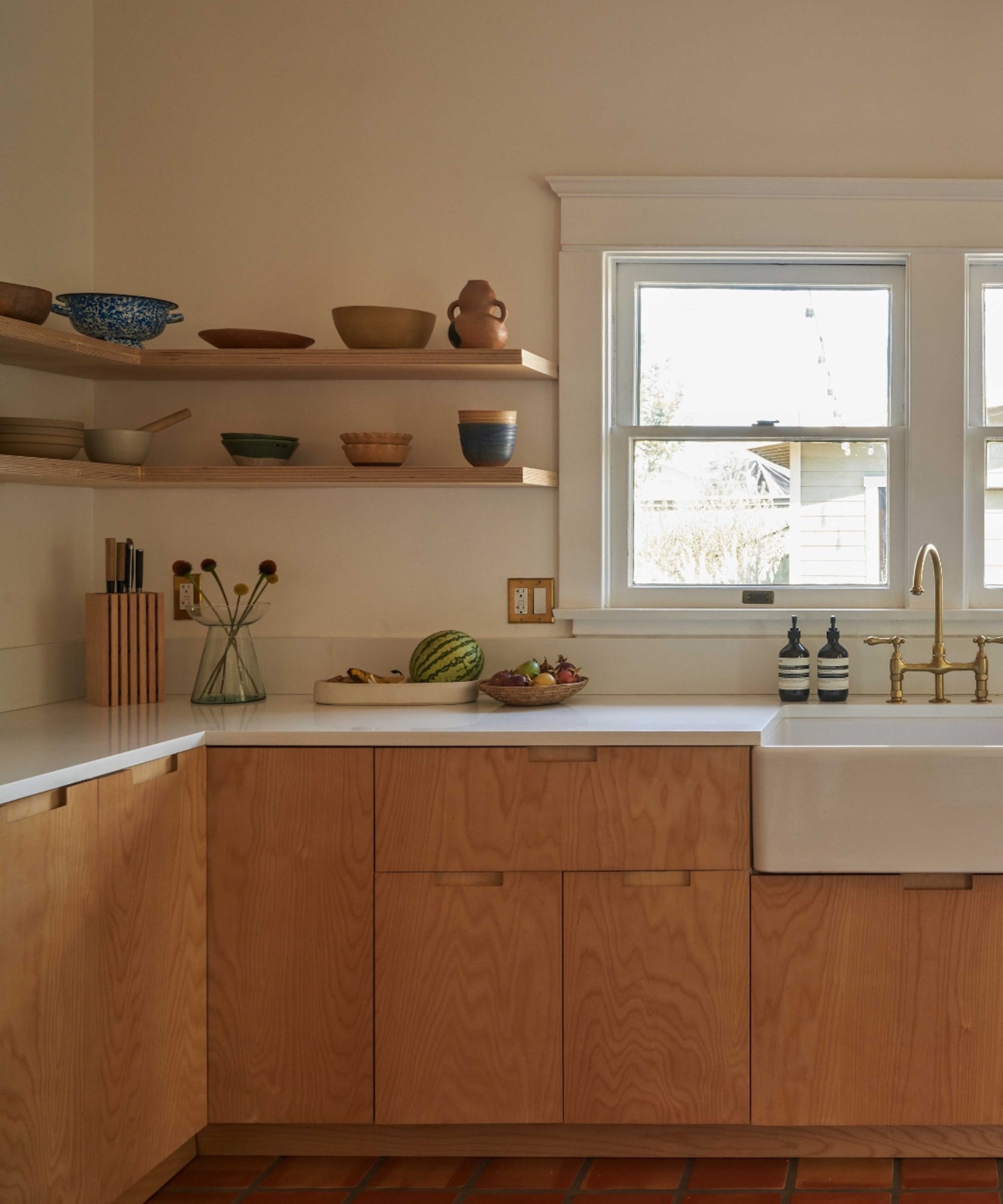 Detail shot of a kitchen sink area with light wood cabinets, white countertops, corner floating shelves, and a brass bridge-style faucet.