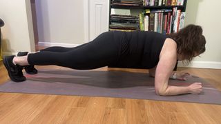 Personal trainer Jennifer Rizzuto performs a plank in her living room on an exercise mat. Her forearms and toes rest on the ground while the rest of her body is elevated off the floor in a straight line. She wears leggings, a vest top, Skechers trainers and has her hair tied back.