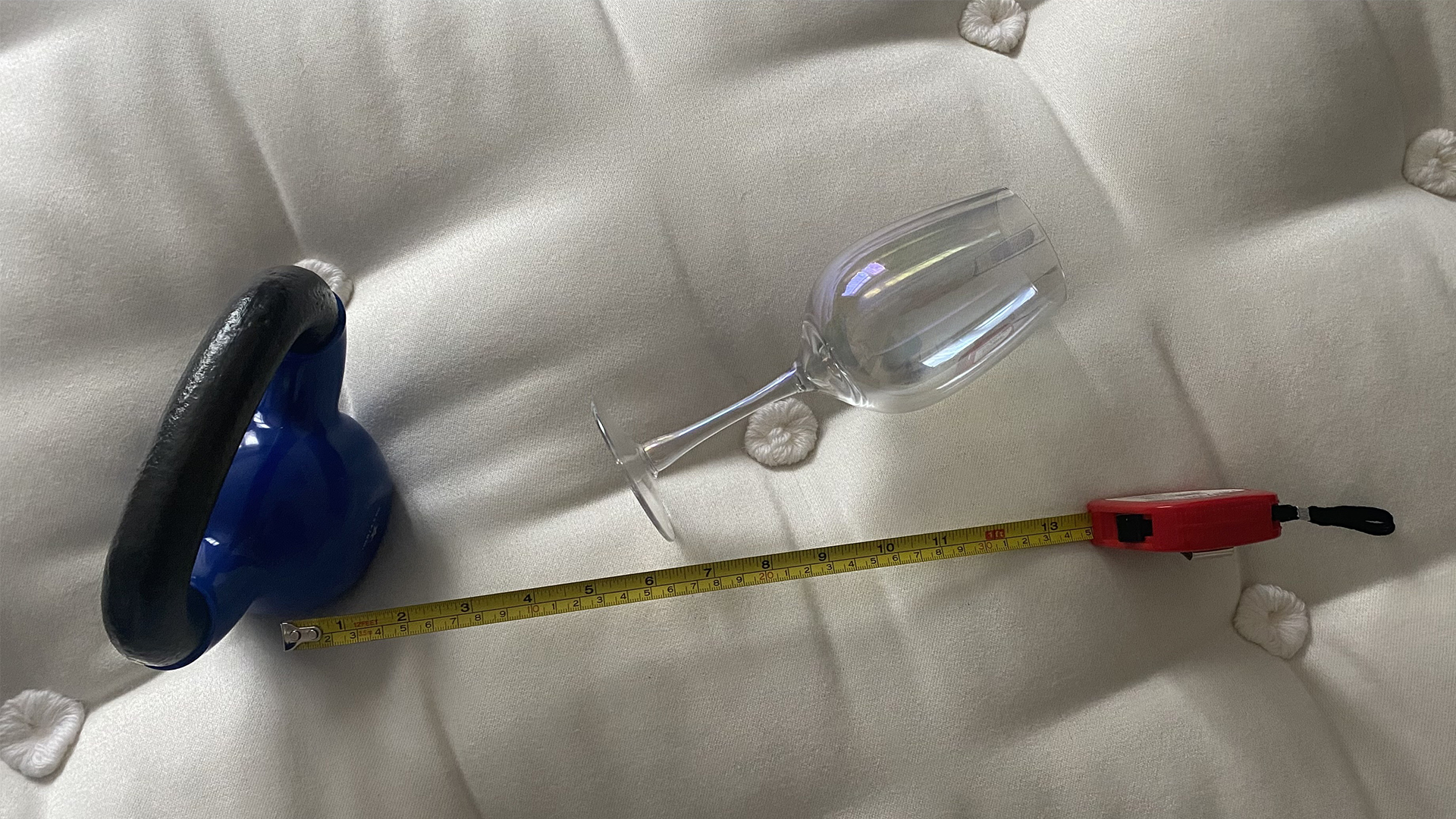 A 4.5kg weight resting on the surface of a Simba Earth Escape mattress, a wine glass has fallen over next to it, anda. tape measure shows there is three inches between the kettlebell and the glass