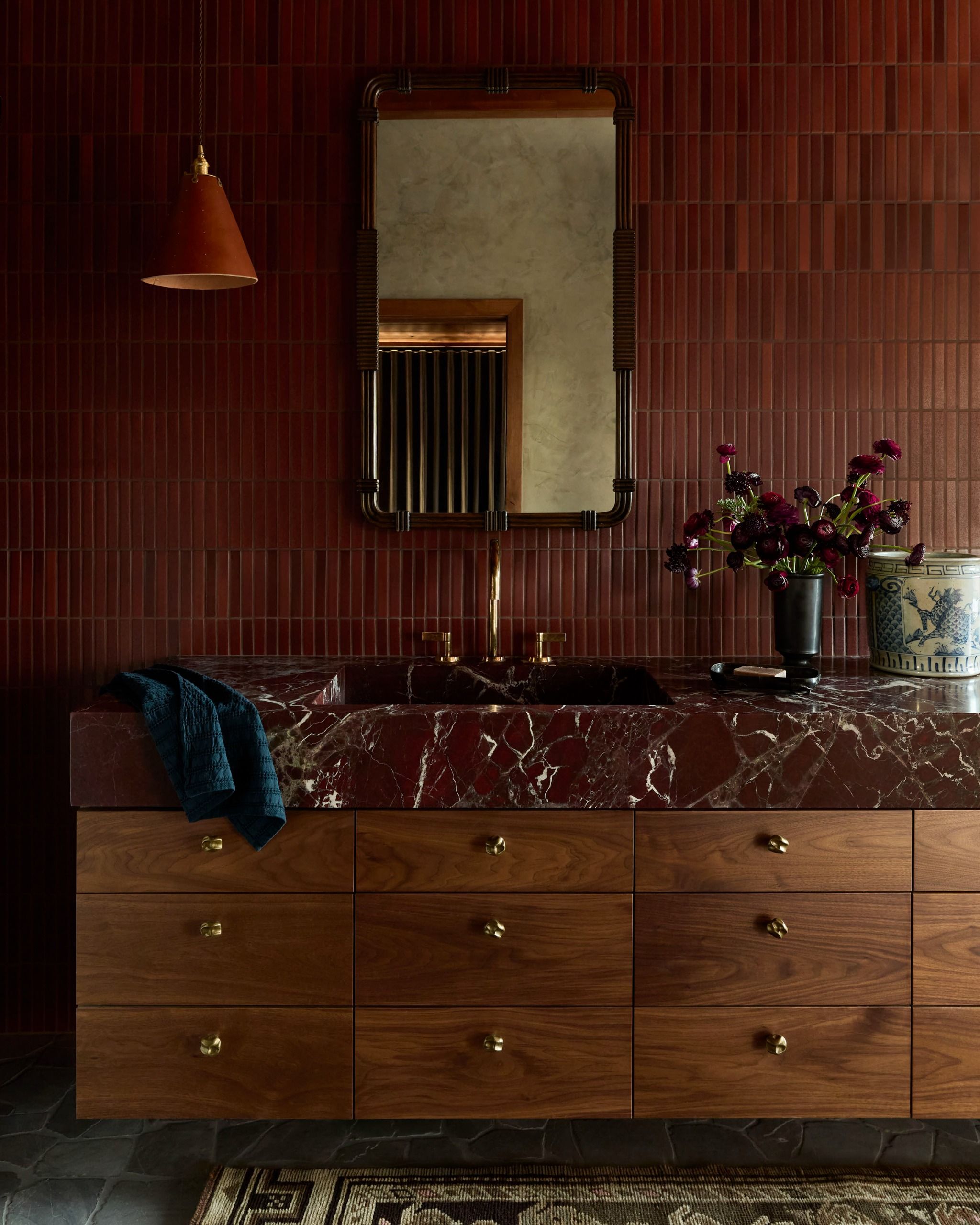 A modern bathroom with a red marble counter on the vanity with wood drawers and brass knobs, thin red tiles are on the wall behind a mirror and red code pendant light with flowers on the vanity
