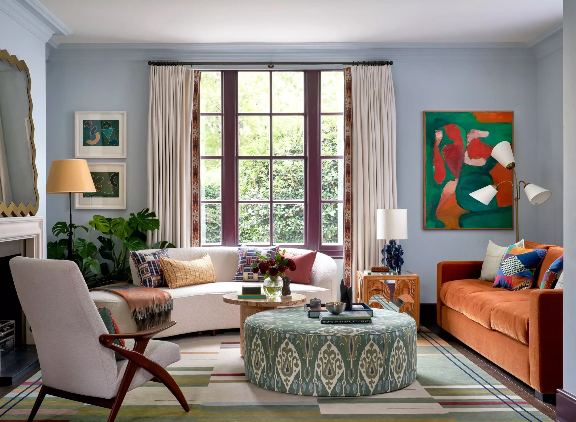 light blue living room with curtains, cream curved sofa, orange velvet sofa, green patterned ottoman coffee table, and beige and timber armchair, with fireplace, lamp, and plant in the corner