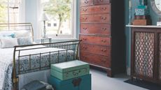 Blue guest bedroom with gold bed, dark wood chest of drawers and cabinet, blue trunks piled at the foot of the bed, and large bay windows.