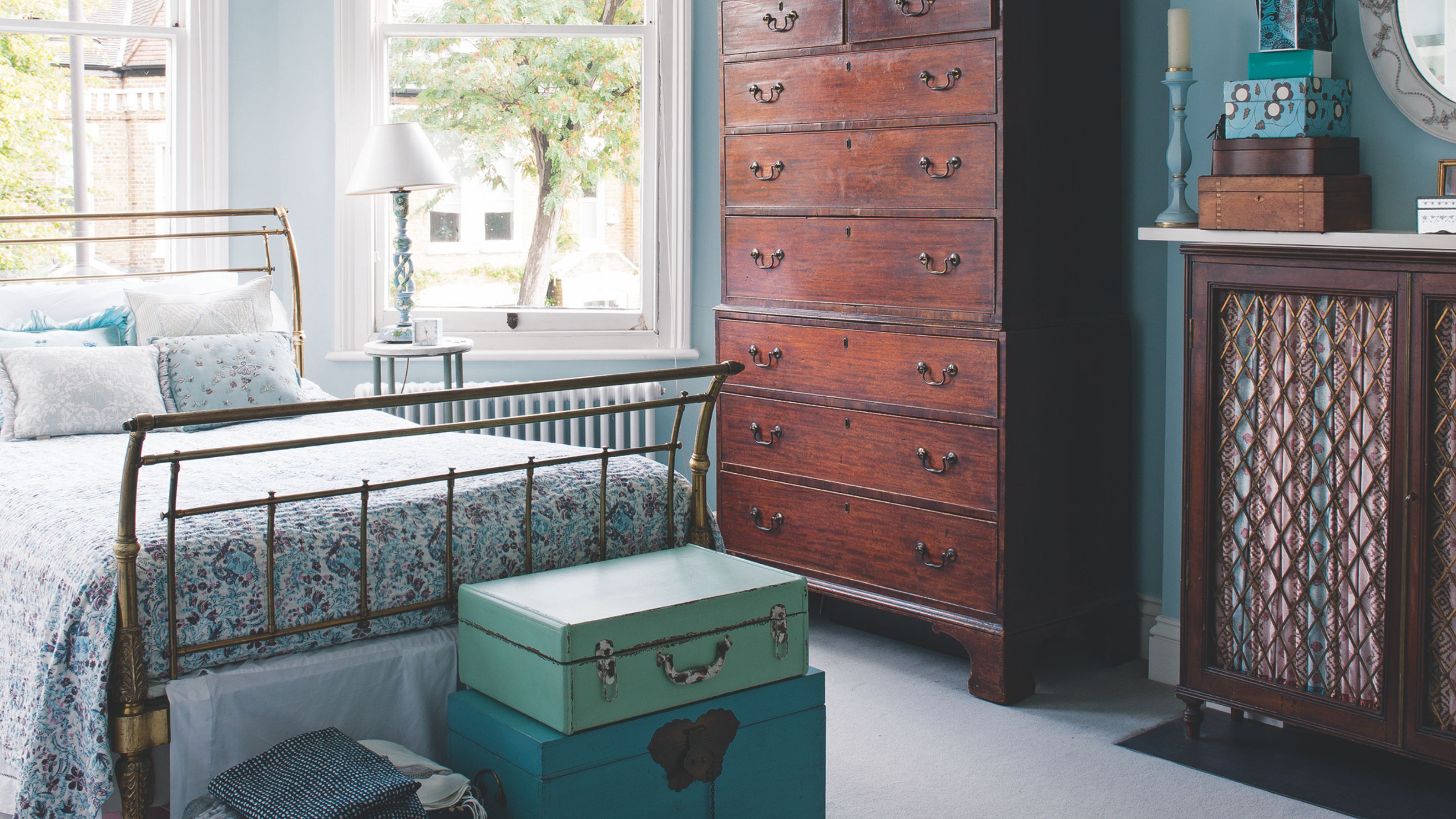 Blue guest bedroom with gold bed, dark wood chest of drawers and cabinet, blue trunks piled at the foot of the bed, and large bay windows.