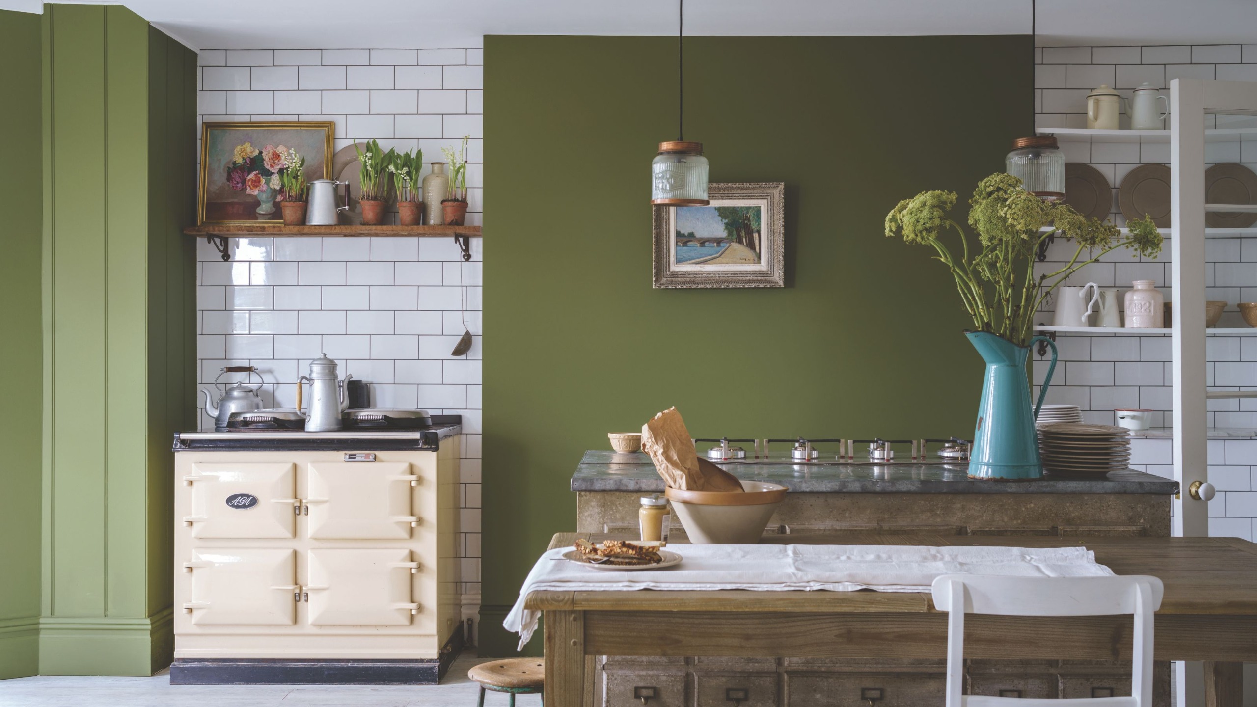 Farmhouse kitchen painted in Bancha by Farrow &amp; Ball, white subway tiles, a cream range cooker, and a wooden dining table with a tablecloth and a blue enamel jug is filled with flowers.