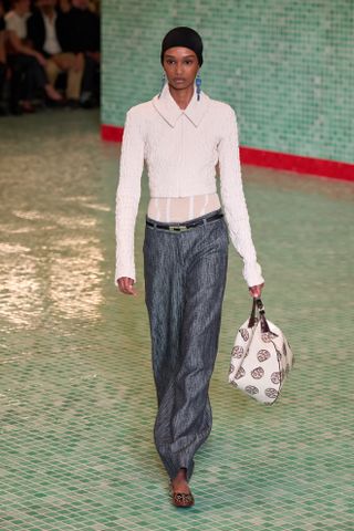 A model walking in the S/S 25 Tory Burch show during NYFW wearing a cream blazer, gray pants, and round toe flats.