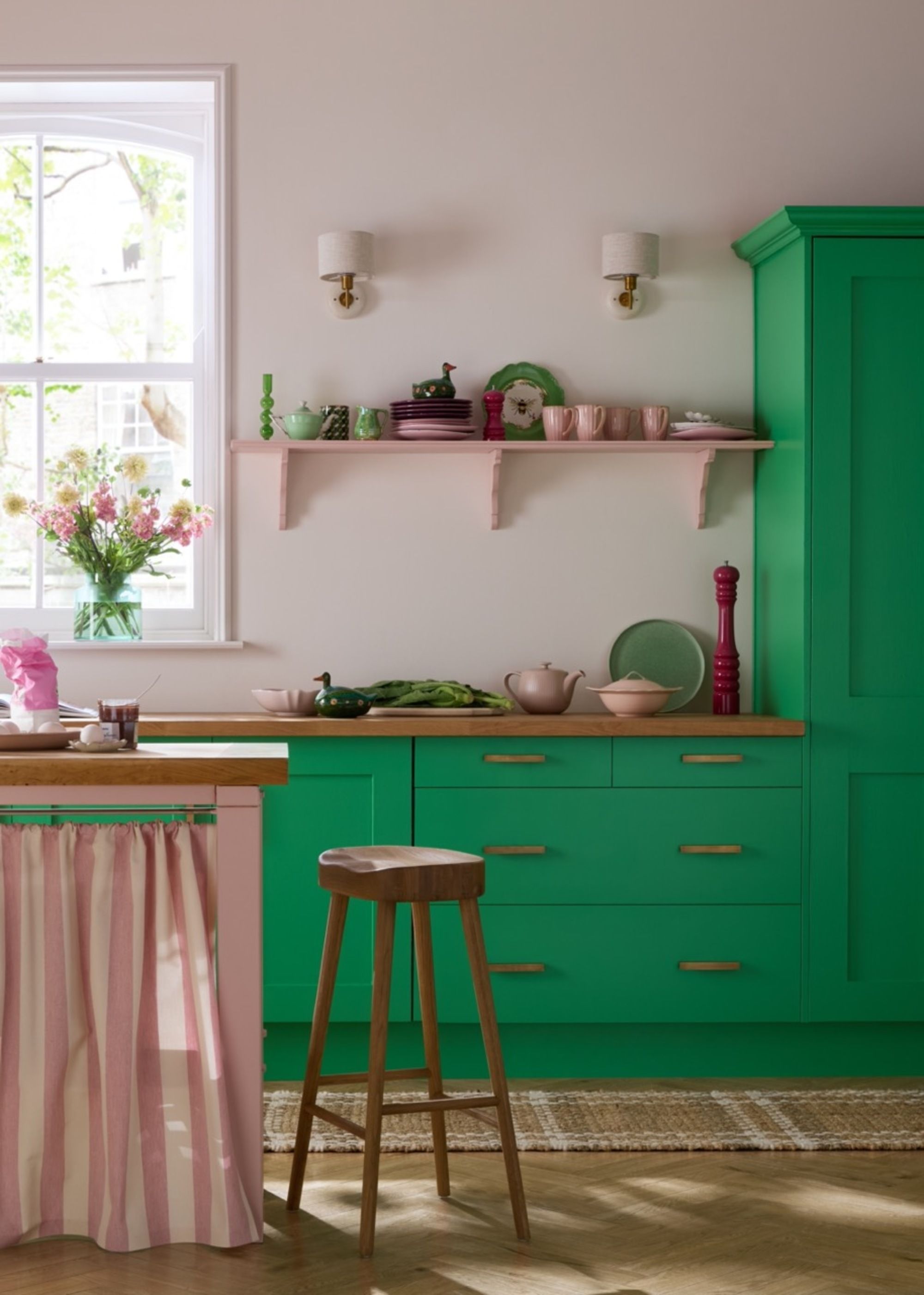 A light pink kitchen with bright Kelly green cabinetry and wooden countertops. There is a pink kitchen island with a pink and white striped cabinet skirt.