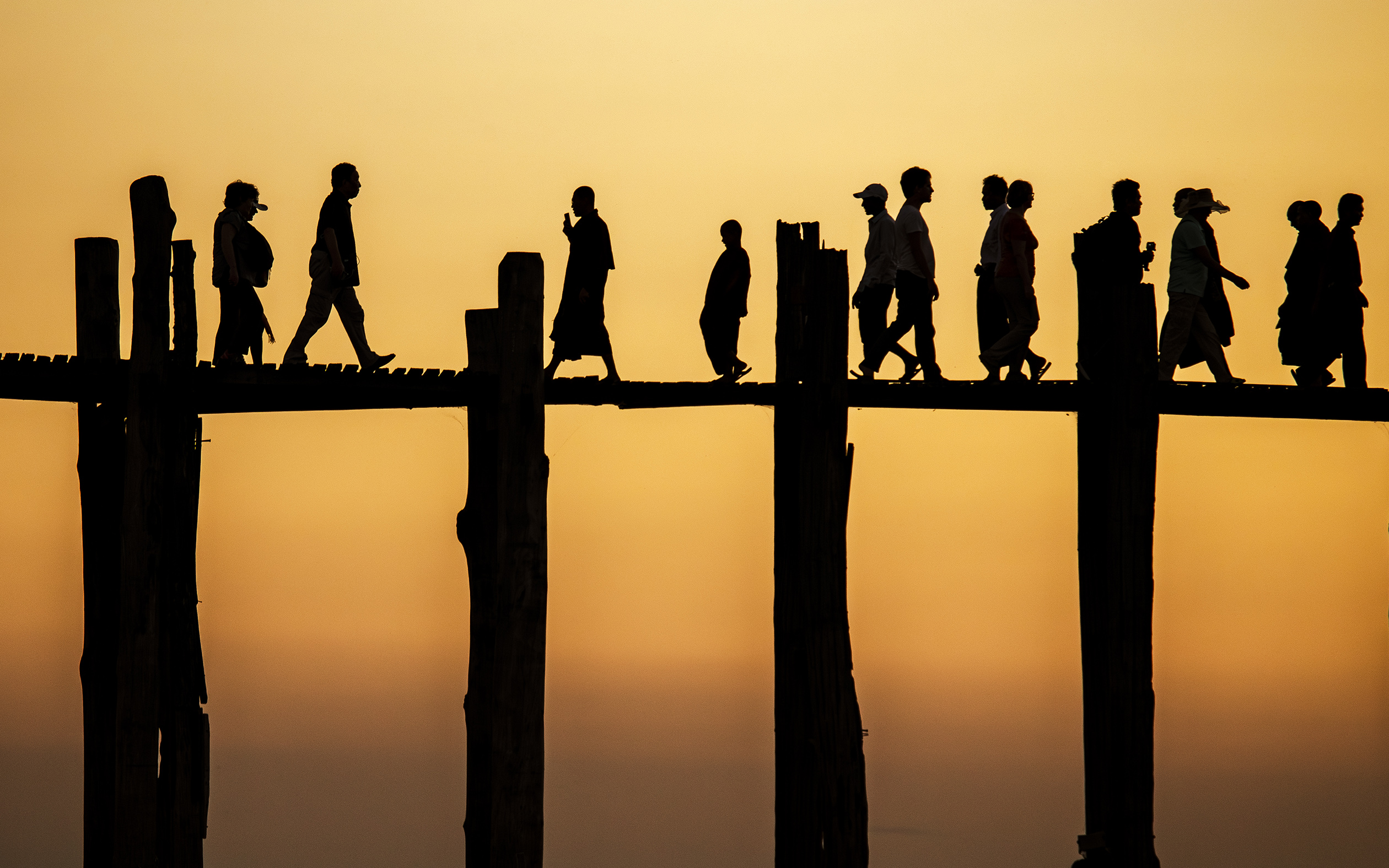 Silhouettes of people walking on a wooden bridge at sunset, set against an orange sky