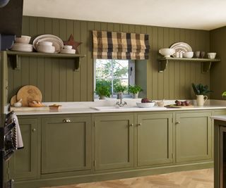 A small kitchen with the cabinets and walls painted olive green, marble countertops, and a checkered blind over the window