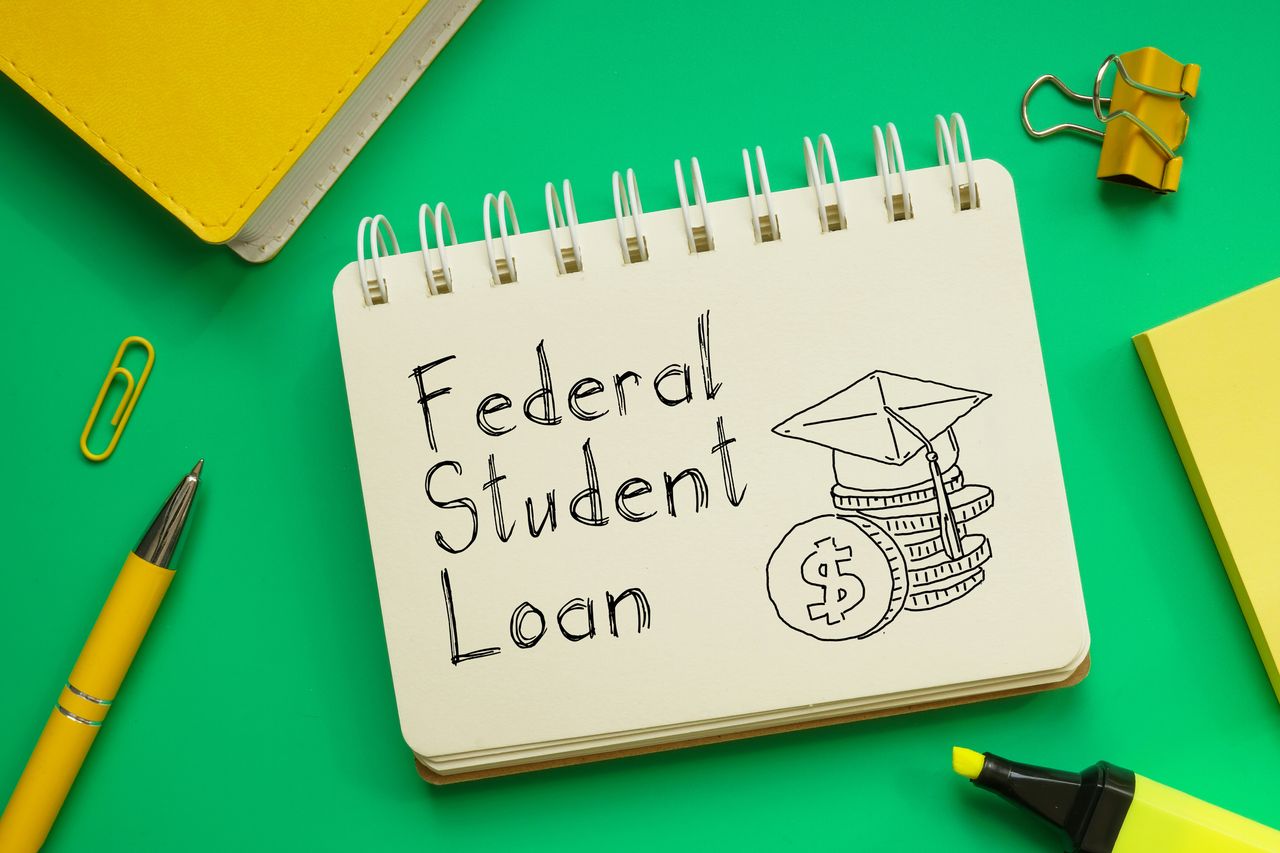 &quot;Federal Student Loan&quot; written on notepad with illustration of graduation cap and coins surrounded by yellow desk accessories against a green background