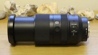 Black Sony E-mount camera lens with zoom extended, placed on a wooden surface with rocks blurred in the background