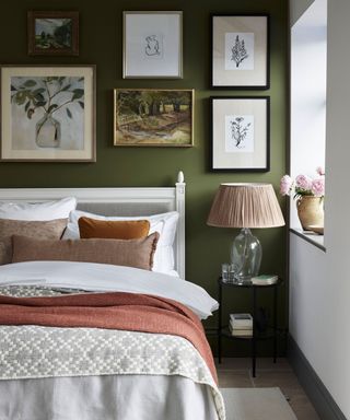 Green painted bedroom with gallery wall above bed, white frame with white linen, cushions and throws in palette of earth colors, metal bedside table with table lamp with glass base, fabric shade, stone flooring, cream rug