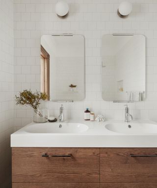 A white tiled bathroom with a white dual sink with a dark wooden contemporary base, two curved mirrors, and two white circular wall sconces above these