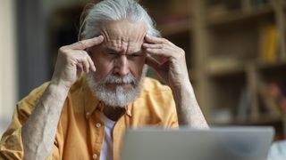 Stressed senior man freelancer working from home, sitting on couch, using laptop, touching his head, having troubles at job, suffering from migraine, headache, copy space, closeup shot