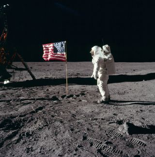 An astronaut stands in front of an American flag on the moon.
