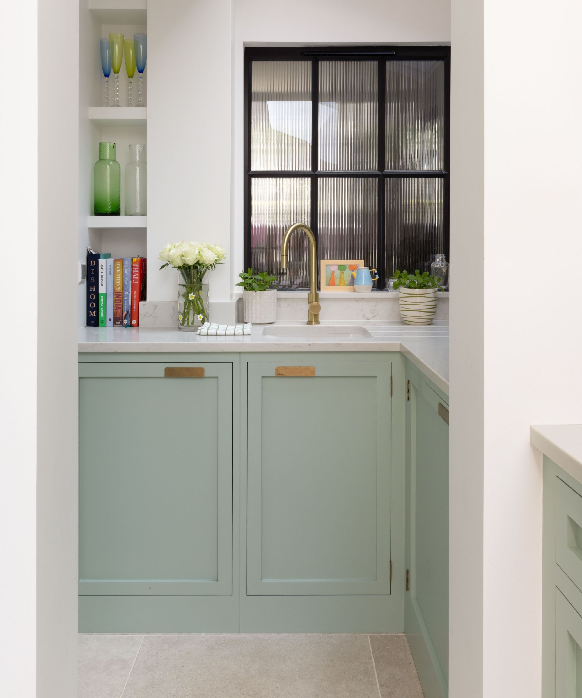 Compact kitchen corner with sage green cabinet doors and brass handles beneath a white marble worktop. A gold tap sits before a black-framed reeded glass window. Open shelves display books and coloured glassware, while white roses and potted herbs soften the space.
