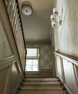 Downwards view of staircase showing patterned walls and window, with a carpet runner on the stairs and a light fixture above.