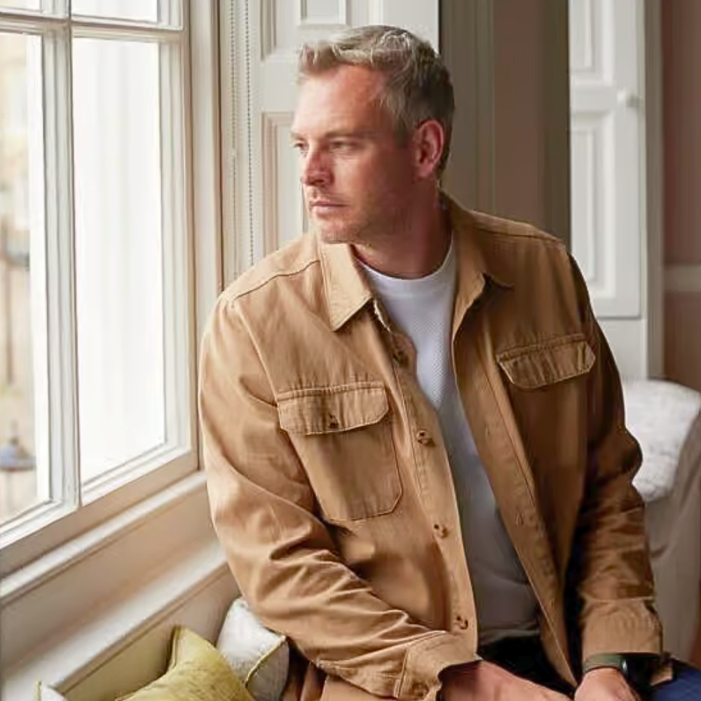 A man with short silver-grey hair sits on a window seat in a bright, naturally lit room, gazing thoughtfully to one side. He wears a tan utility overshirt over a white t-shirt. A cushion and armchair are partially visible beside him.