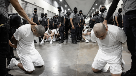 Two men wearing all white kneel on a concrete floor, held by prison guards. Behind them, prisoners are sitting on the group with their heads down, surrounded by masked guards.