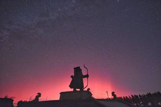 A red sky rises over a stone figure holding a bow and arrow