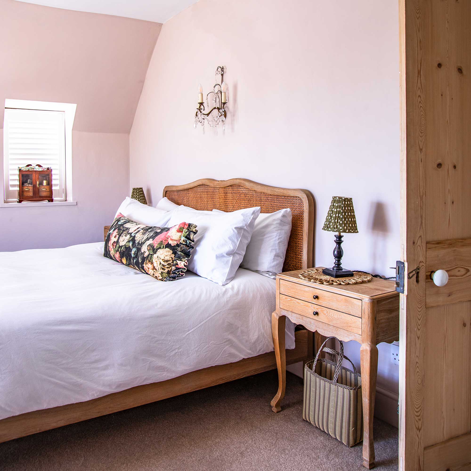 Pale pink painted bedroom with a wooden and rattan bed, a wooden bedside table, neutral bedding
